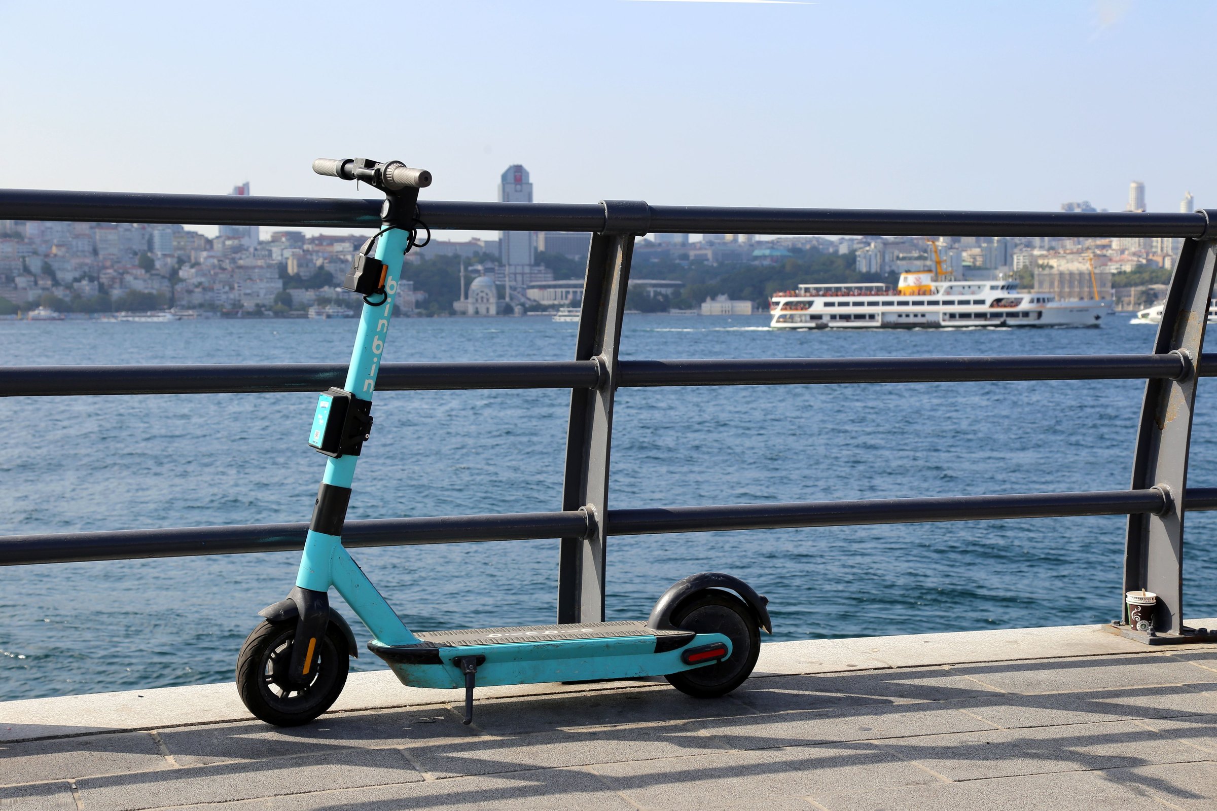 Trottinette électrique turquoise stationnée contre une rambarde noire, ville floue en arrière-plan.